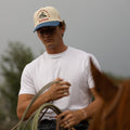 Man wearing an Alpine Days Rodeo cap and holding a lasso, standing next to a horse with a blurred background