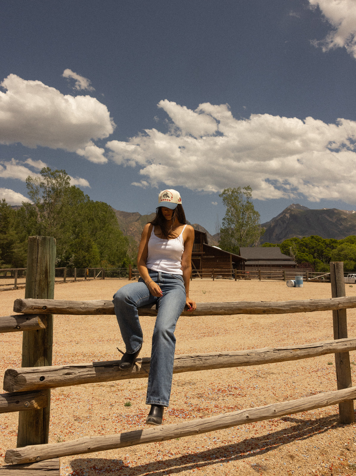 Woman in a Loud Face Supply rodeo cap sitting on a corral fence with mountains behind her